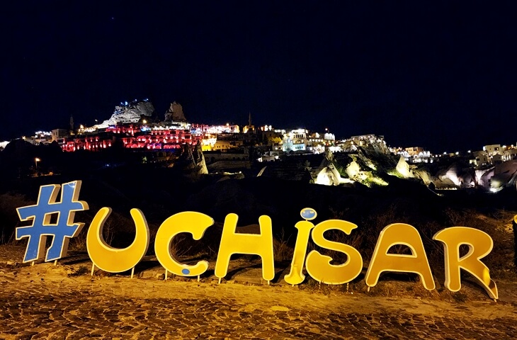 Gece ışıklarıyla aydınlatılmış Uçhisar Kalesi ve ön planda büyük #Uchisar yazısı